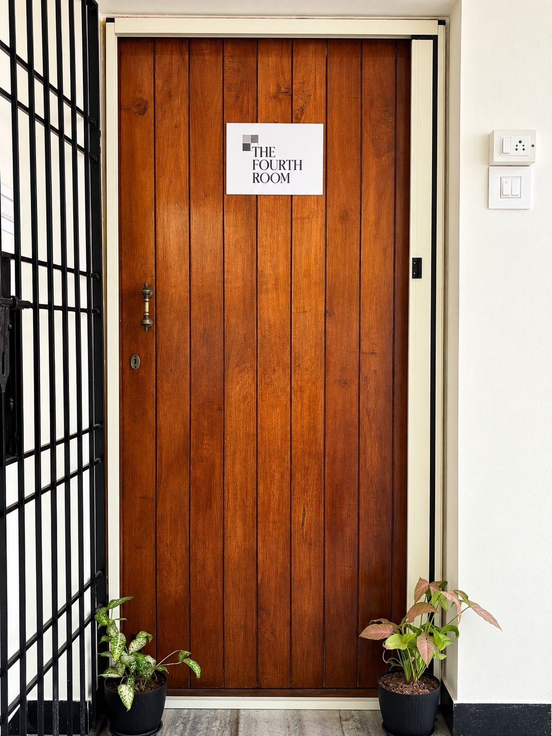 The wooden front door of The Fourth Room with its sign, a black iron grille on one side and potted plants on the floor.