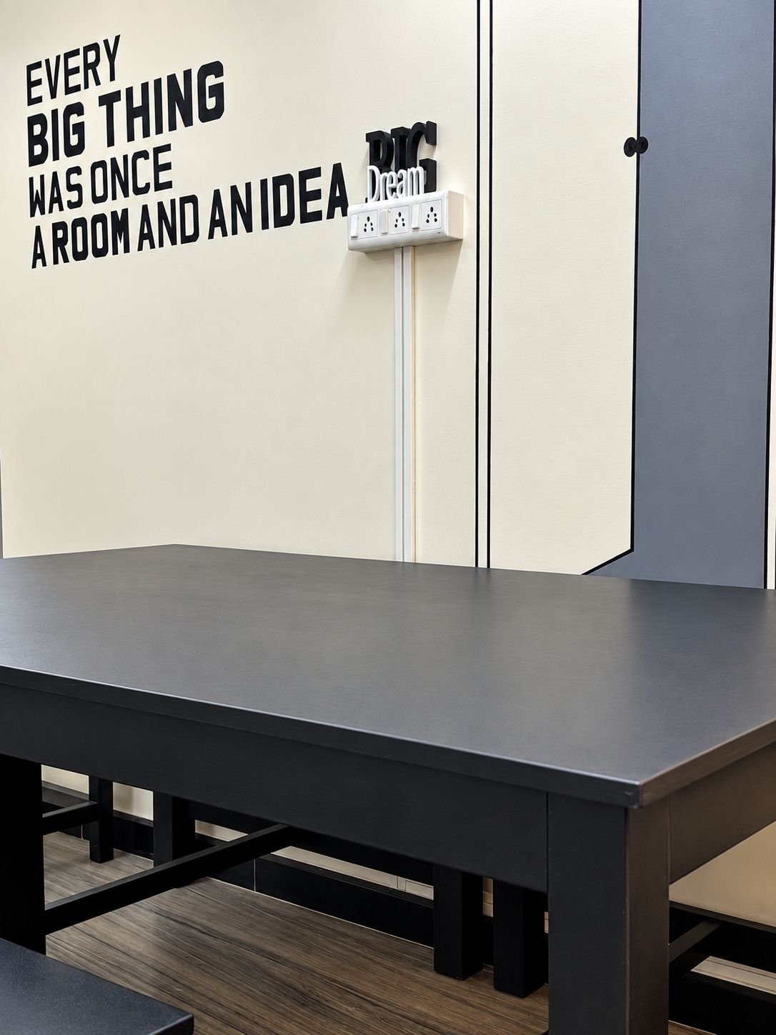 A black table in front of a cream wall painted with the words 'Every big thing was once a room and an idea', with plug points on the wall.