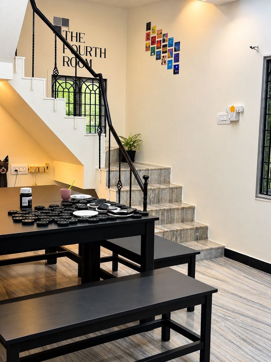 The ground-floor room at The Fourth Room: black tables and benches set with craft supplies, the staircase up to the first floor, the painted sign 'The Fourth Room' and a cluster of small colourful paintings on the wall.