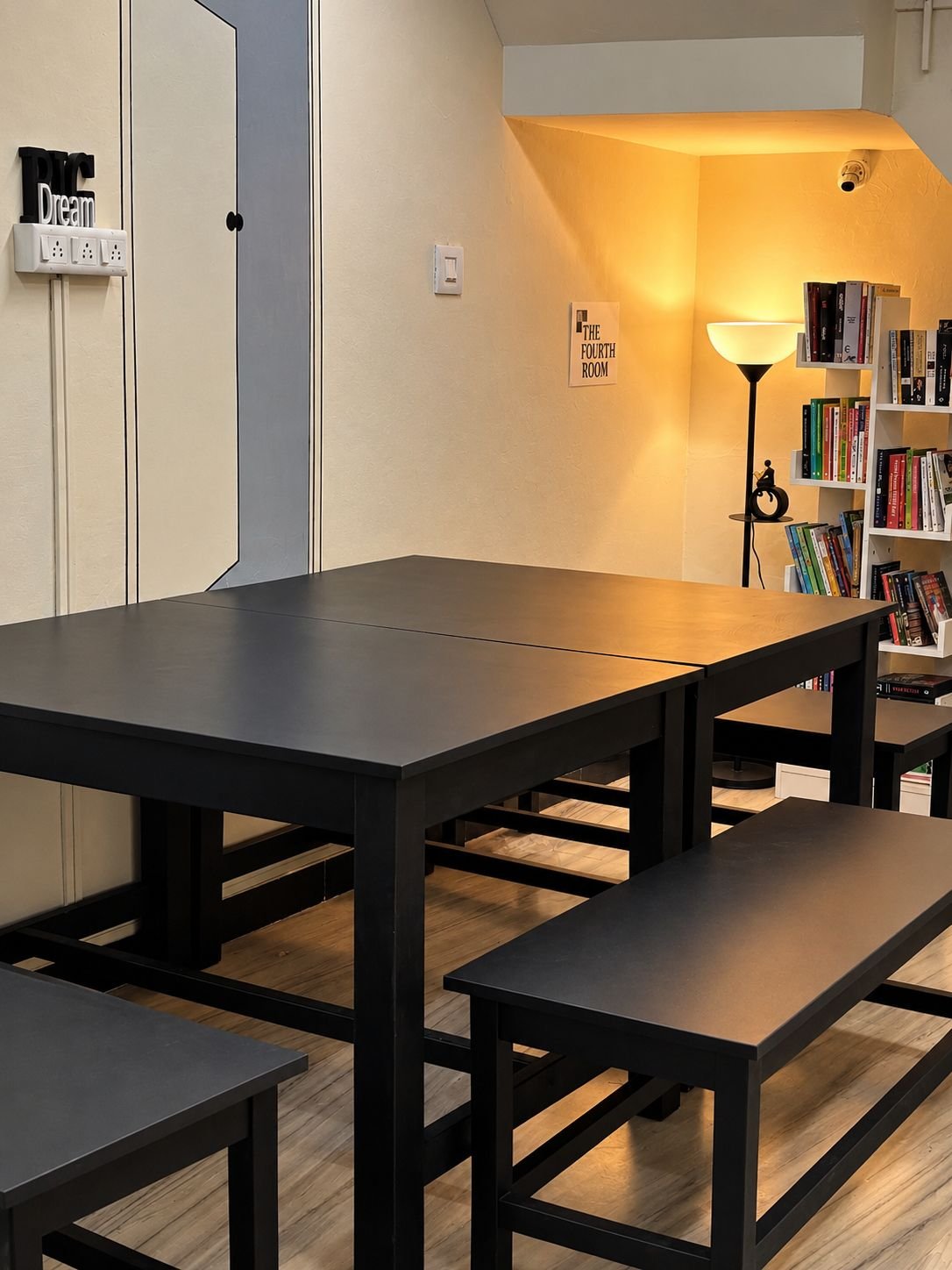 Black tables and benches in The Fourth Room, with the lamp-lit reading nook and bookshelf in the corner under the stairs.